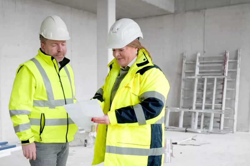 Imagebild Bauleitung Ein Bauleiter und eine Bauleiterin in Gelben Jacken und mit Baustellenhelmen schauen zusammen auf einen Bauplan